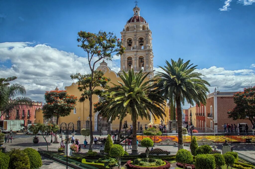 Événements au Mexique en mars 2026 : que faire et où A vibrant Mexican plaza featuring a historic yellow church tower surrounded by lush palm trees and traditional colonial architecture