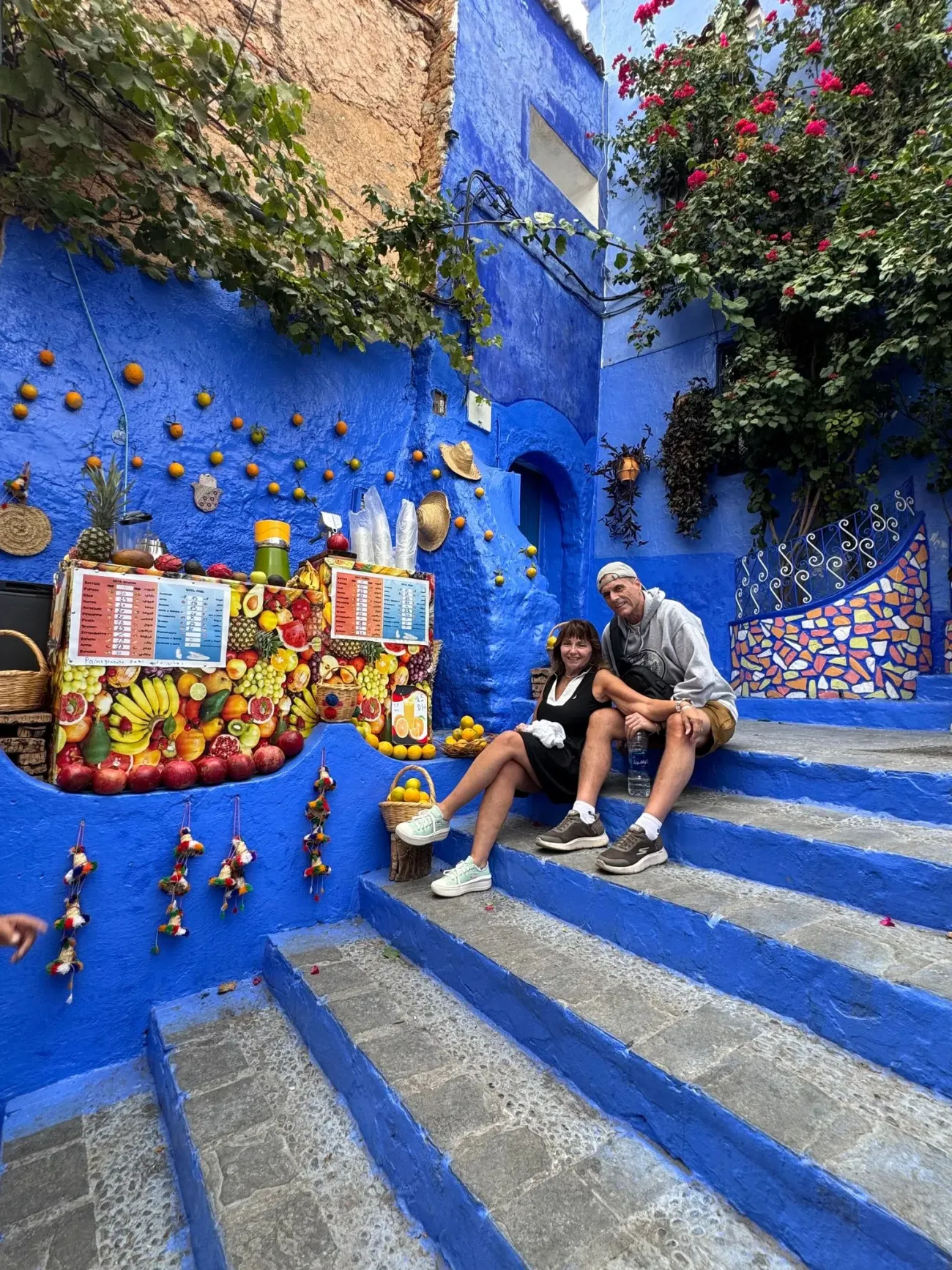 Marches et murs peints en bleu à Chefchaouen, au Maroc, avec deux voyageurs assis dans les escaliers à côté d'un étal de fruits et de jus colorés.