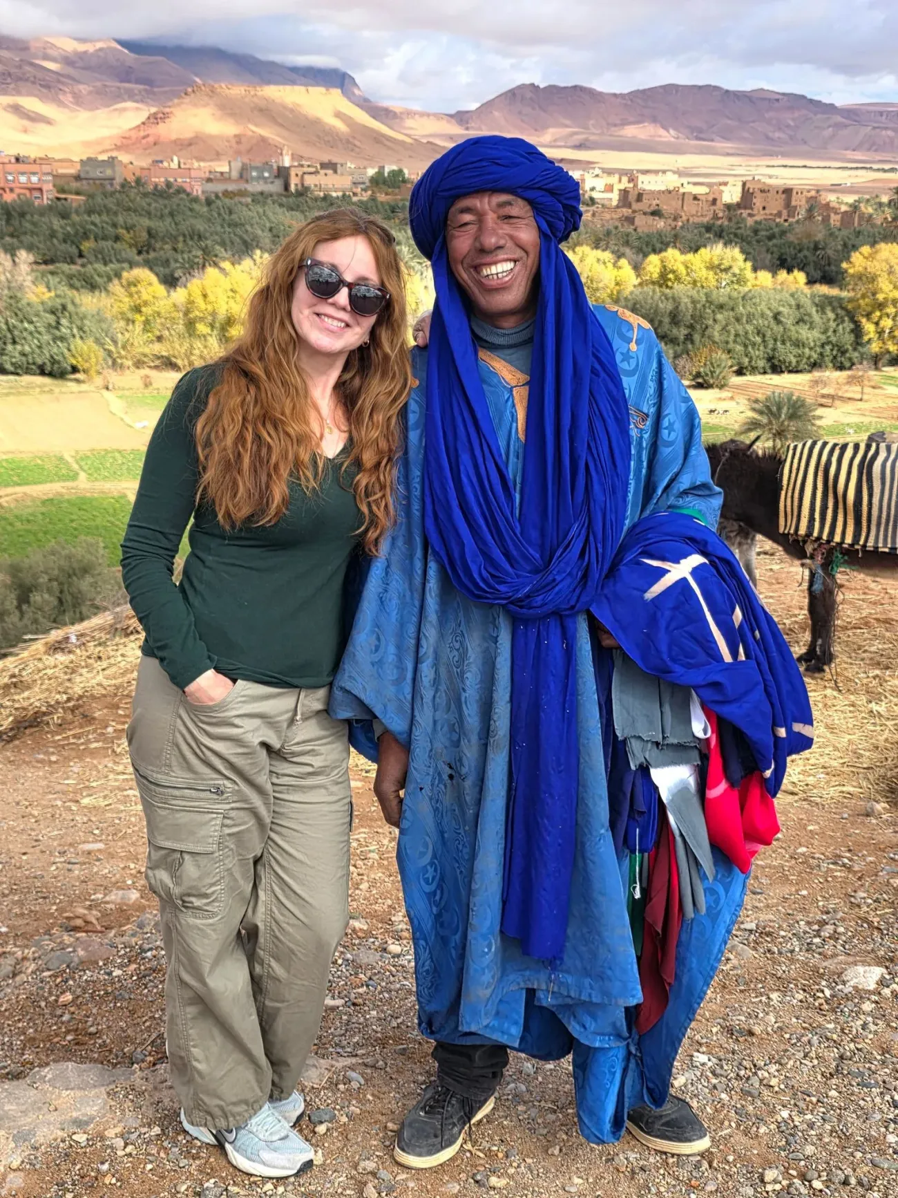 Homme en robe bleue vibrante debout avec un voyageur devant une oasis de palmiers et des kasbahs dans la vallée du Draa, au Maroc.