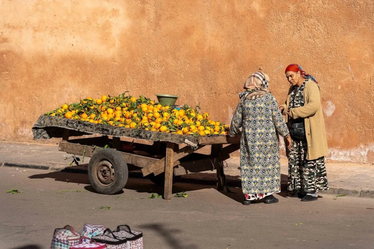 Chariot de rue en bois rempli d'oranges vives à côté de deux femmes parlant contre un mur ocre au Maroc.
