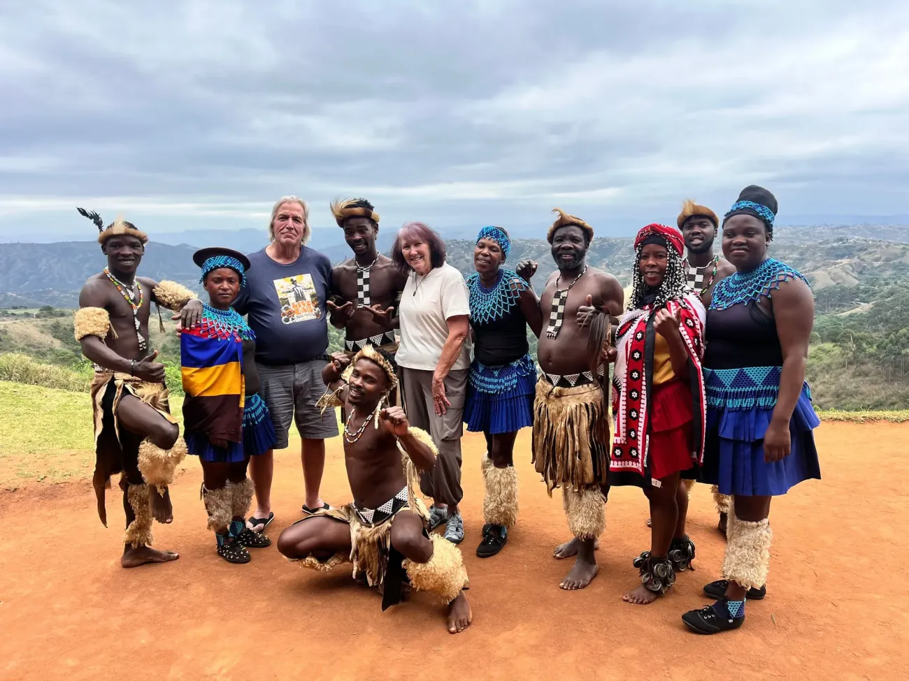 Danseurs locaux et deux touristes posant ensemble sur un terrain en terre battue avec des collines en arrière-plan, Afrique du Sud.